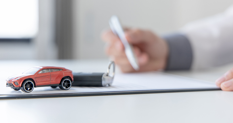 Small car on a table with keys nearby. Hand with a pen in the background.