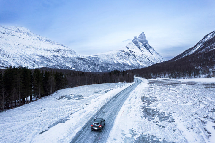 Aerial view of car driving down a snowy road with forest on the sides and mountains ahead. Daytime driving. Winter Scene.